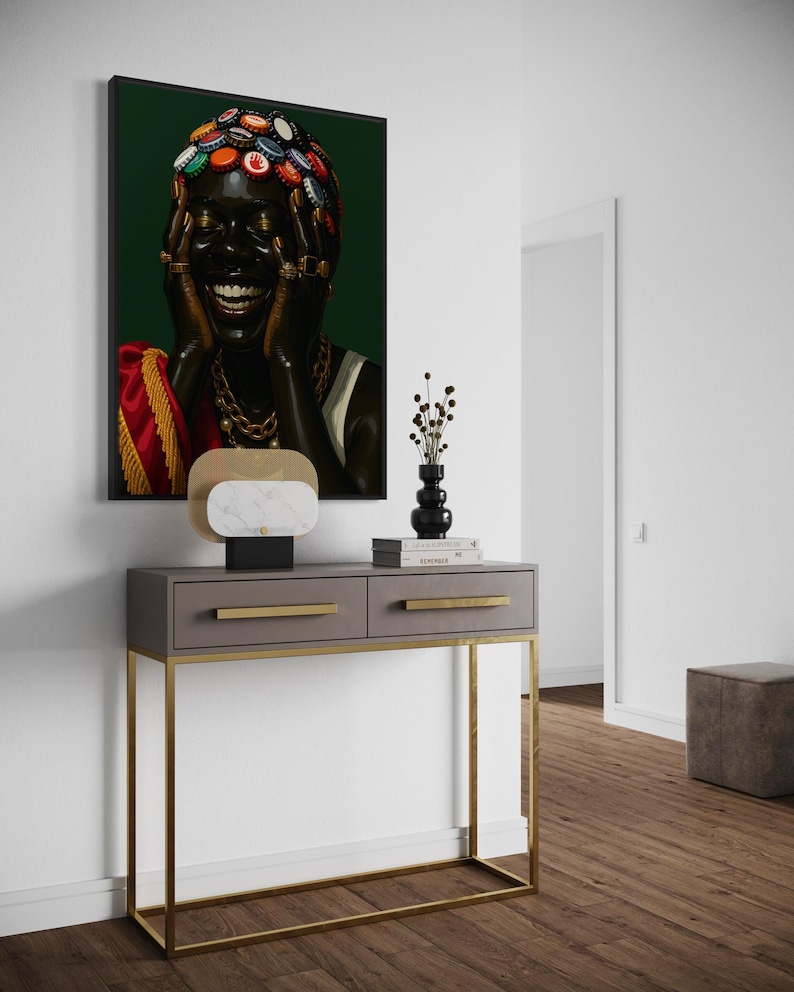 May include: A framed portrait of a person with dark skin, adorned with bottle caps on their head and gold jewelry, laughing. The artwork hangs above a gray console table with gold legs and drawer pulls, complemented by a decorative lamp and vase.