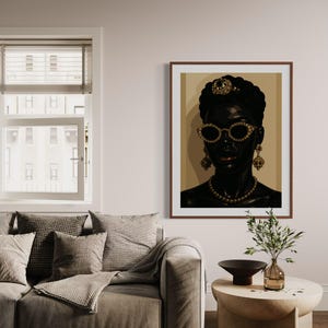 May include: A framed portrait of a person with dark skin, wearing gold-rimmed sunglasses, a jeweled headpiece, and earrings. The portrait is displayed in a living room setting with a sofa, pillows, and a side table.