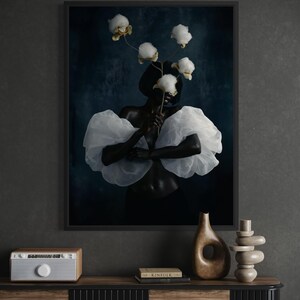 May include: A framed art print featuring a person with dark skin wearing a white, ruffled top and holding cotton blossoms. The background is a dark blue, and the print is displayed above a wooden cabinet with decorative items.