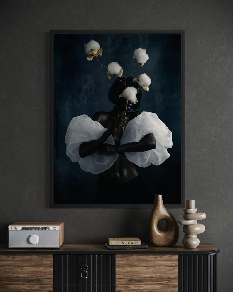 May include: A framed art print featuring a person with dark skin wearing a white, ruffled top and holding cotton blossoms. The background is a dark blue, and the print is displayed above a wooden cabinet with decorative items.