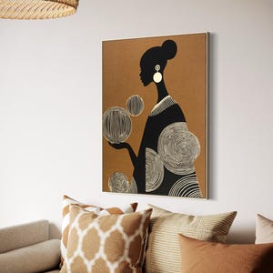 May include: A framed art print featuring a black silhouette of a person with a bun, wearing large white earrings, and a black dress. The background is a warm brown, and the person is holding three white and black circular designs. The artwork is displayed above a sofa with decorative pillows.