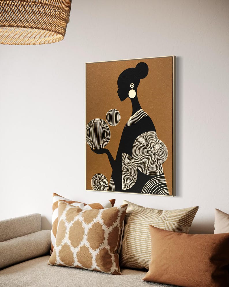 May include: A framed art print featuring a black silhouette of a person with a bun, wearing large white earrings, and a black dress. The background is a warm brown, and the person is holding three white and black circular designs. The artwork is displayed above a sofa with decorative pillows.