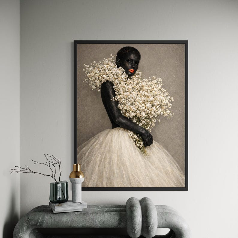 May include: A framed art print featuring a person with dark skin, wearing a white tulle skirt and a collar of white flowers. The person holds a bouquet of white flowers and has red lipstick. The background is a neutral tone.