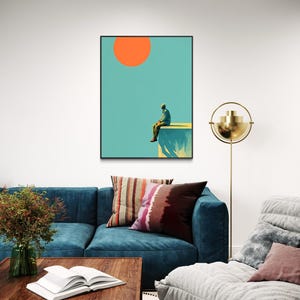 May include: A framed art print featuring a person sitting on a ledge under a large orange sun against a teal sky. The artwork is displayed above a blue sofa with decorative pillows, a wooden coffee table, and a gold floor lamp.