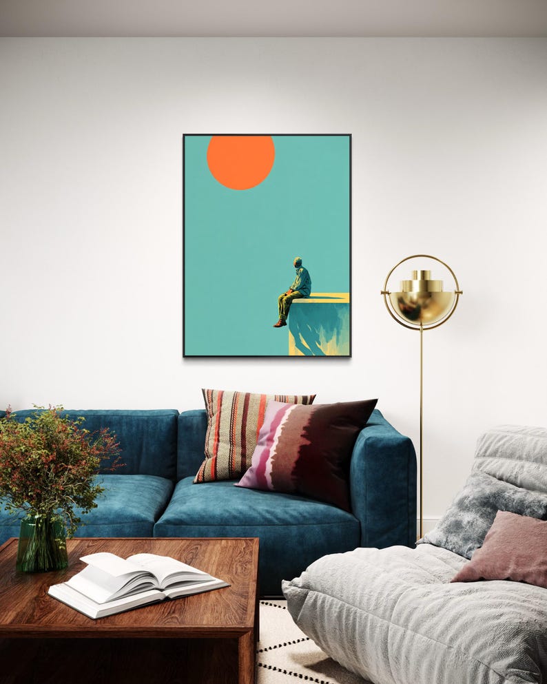 May include: A framed art print featuring a person sitting on a ledge under a large orange sun against a teal sky. The artwork is displayed above a blue sofa with decorative pillows, a wooden coffee table, and a gold floor lamp.