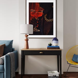 May include: A framed art print depicts a person holding a glass of red liquid, with a gold chain necklace. The artwork is set against a red backdrop. A wooden table with a lamp, books, and a vase is in front of a blue sofa.