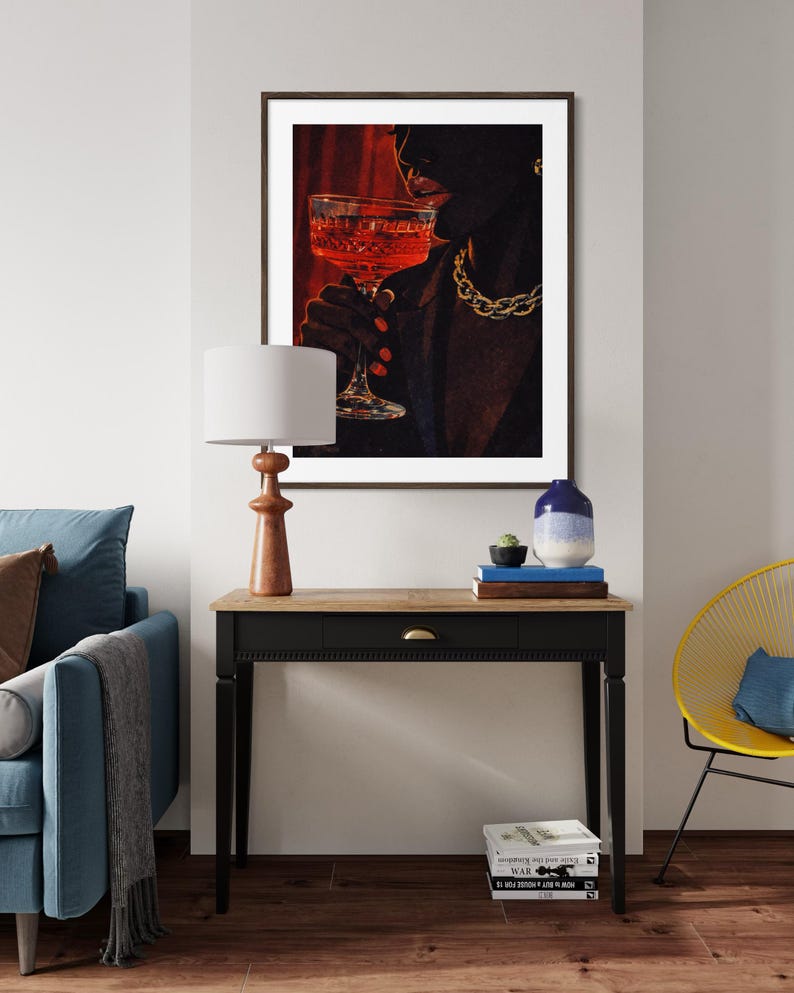 May include: A framed art print depicts a person holding a glass of red liquid, with a gold chain necklace. The artwork is set against a red backdrop. A wooden table with a lamp, books, and a vase is in front of a blue sofa.
