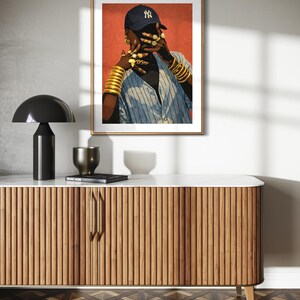 May include: A framed art print featuring a person wearing a blue baseball cap with a NY logo, gold jewelry, and a striped shirt. The artwork is displayed above a wooden cabinet with a marble top, a black lamp, and decorative items.