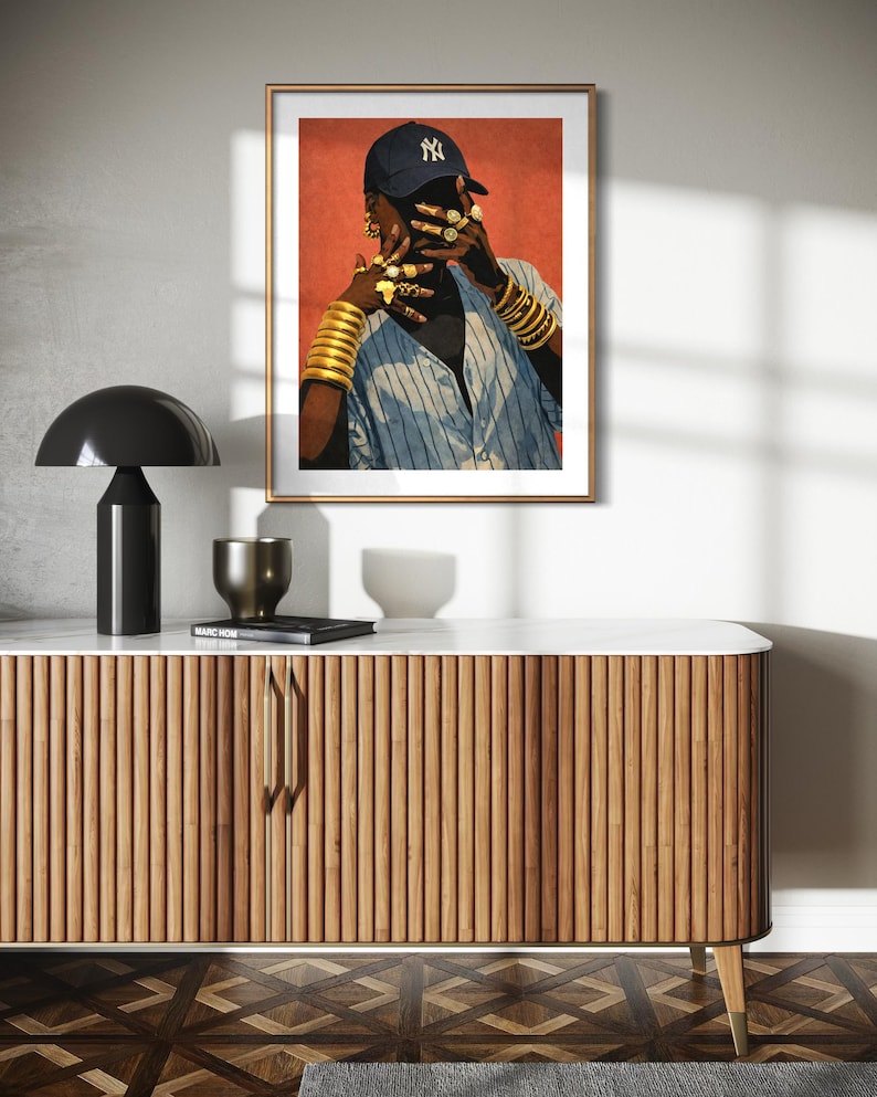 May include: A framed art print featuring a person wearing a blue baseball cap with a NY logo, gold jewelry, and a striped shirt. The artwork is displayed above a wooden cabinet with a marble top, a black lamp, and decorative items.