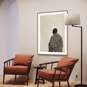 May include: A room interior with two wooden armchairs with terracotta cushions, a framed artwork with a figure in black and white stripes, and a black floor lamp. The walls are light-coloured and the floor is wooden.