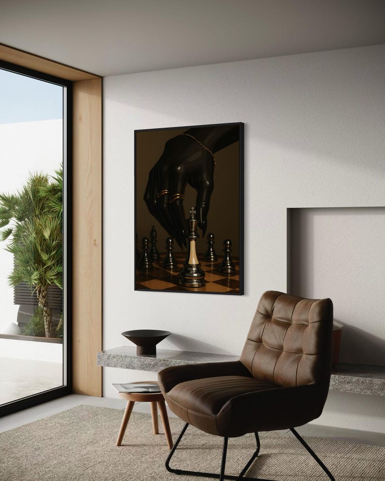 Op de afbeelding: Een ingelijst kunstwerk dat een zwarte hand met gouden sieraden afbeeldt die een schaakstuk verplaatst. De schaakstukken staan op een schaakbord. Het kunstwerk wordt getoond in een moderne kamer met een bruine leren stoel en een kleine houten kruk.