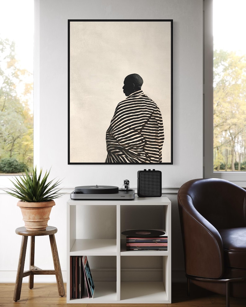May include: A monochrome art print shows a person in profile, wearing a striped robe. The artwork is above a white cube shelf with vinyl records and a record player. A potted plant sits on a wooden stool, next to a window.