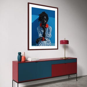 May include: A framed portrait of a person wearing red jewellery and a striped shirt hangs above a red and blue cabinet. A red lamp with a gold base sits on the cabinet. A colourful striped rug is on the floor.