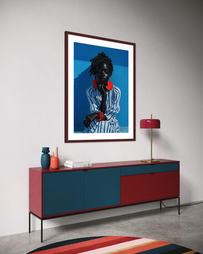 May include: A framed portrait of a person wearing red jewellery and a striped shirt hangs above a red and blue cabinet. A red lamp with a gold base sits on the cabinet. A colourful striped rug is on the floor.