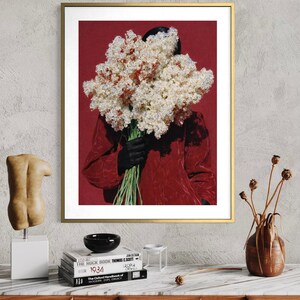 May include: A framed art print featuring a person holding a large bouquet of white and red flowers against a red background. The person is wearing a red jacket and black gloves. The artwork is displayed on a wall above a wooden cabinet.
