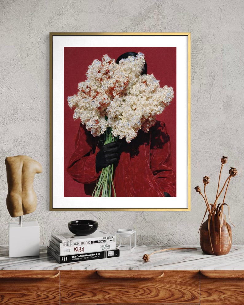 May include: A framed art print featuring a person holding a large bouquet of white and red flowers against a red background. The person is wearing a red jacket and black gloves. The artwork is displayed on a wall above a wooden cabinet.