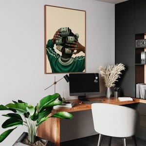 May include: Framed art print featuring a person with dollar bills styled as hair rollers. The person is wearing a green and white track suit. The artwork is displayed in a modern office setting with a wooden desk, computer, and a white chair.