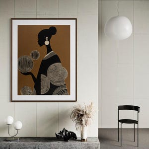 May include: A framed art print featuring a black and white silhouette of a woman holding circular designs. The artwork is set against a brown background with a white border. The print is displayed in a modern interior setting.