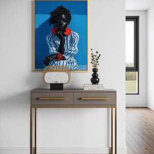 May include: A framed portrait featuring a person with dark skin, wearing a blue and white striped dress and red jewellery. The portrait hangs above a grey console table with gold accents. A black vase and decorative object sit on the table.