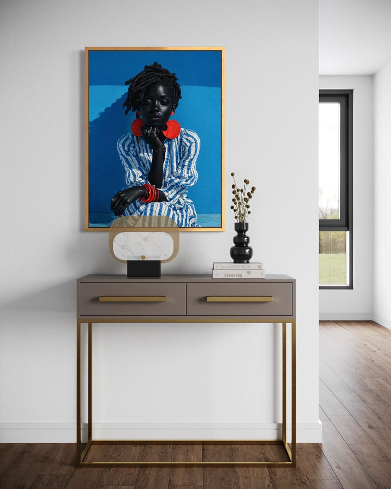May include: A framed portrait featuring a person with dark skin, wearing a blue and white striped dress and red jewellery. The portrait hangs above a grey console table with gold accents. A black vase and decorative object sit on the table.