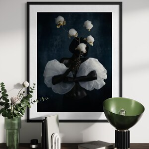 May include: A framed art print featuring a person with dark skin holding cotton blossoms, wearing a white ruffled top. The artwork is set against a dark blue background. A green lamp and decorative items are on a wooden shelf.