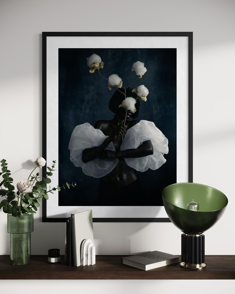 May include: A framed art print featuring a person with dark skin holding cotton blossoms, wearing a white ruffled top. The artwork is set against a dark blue background. A green lamp and decorative items are on a wooden shelf.