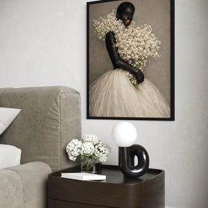 May include: A framed wall art print featuring a person with dark skin and bright orange lips, wearing a voluminous white tulle skirt and holding a large bouquet of white baby's breath flowers against a neutral gray background.