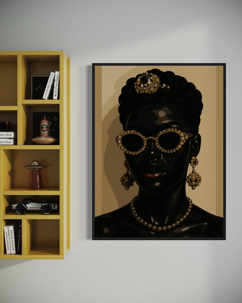 May include: A framed art print featuring a stylized portrait of a person with dark skin. The person is adorned with gold-colored jewelry, including a necklace, earrings, sunglasses, and a headpiece. The background is a neutral beige.