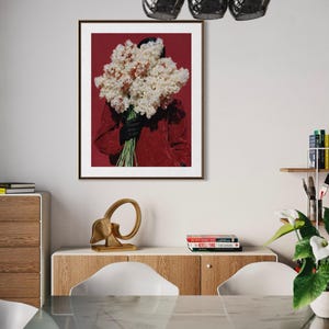 May include: A framed art print featuring a person holding a large bouquet of white and pink flowers against a deep red background. The person is wearing a red top and black gloves. The artwork is displayed in a modern interior setting.