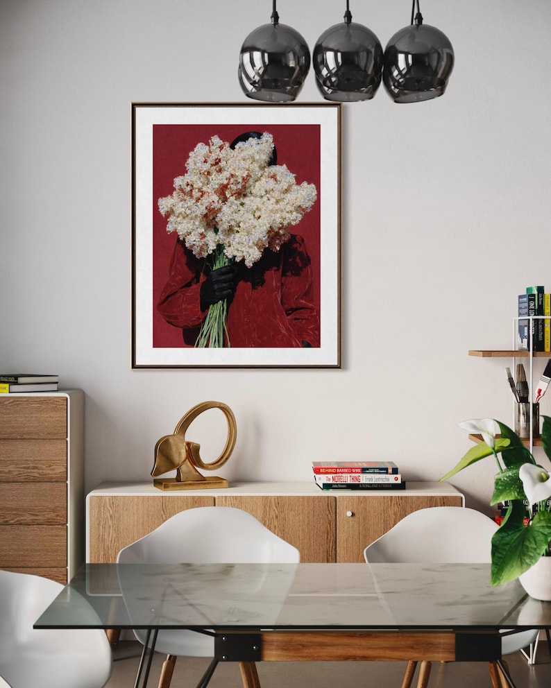 May include: A framed art print featuring a person holding a large bouquet of white and pink flowers against a deep red background. The person is wearing a red top and black gloves. The artwork is displayed in a modern interior setting.