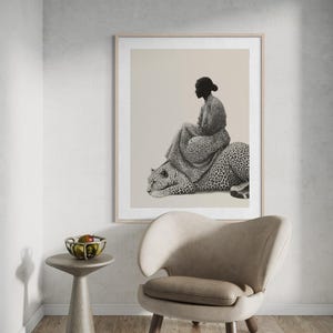 May include: A framed black and white print depicts a person seated on a leopard. The person is wearing a patterned dress. The artwork is displayed above a modern chair and a small side table with a bowl of fruit.