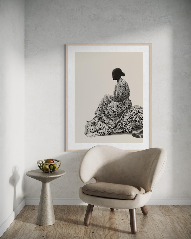May include: A framed black and white print depicts a person seated on a leopard. The person is wearing a patterned dress. The artwork is displayed above a modern chair and a small side table with a bowl of fruit.
