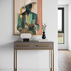 May include: An abstract painting of a person in a hat and suit, with green and beige tones, hangs above a gray console table with gold accents. A decorative object with a marble top and a black vase with dried flowers sit on the table. The room has hardwood floors.
