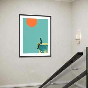 May include: Framed art print featuring a minimalist illustration. A person sits on the edge of a cliff, with a large orange sun in a teal sky. The artwork is in a black frame, suitable for home decor.