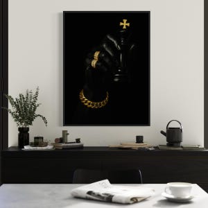 May include: A framed art print featuring a black chess king with a gold cross on top, held by a gloved hand adorned with gold rings and a gold chain bracelet. The background is solid black, creating a striking contrast.
