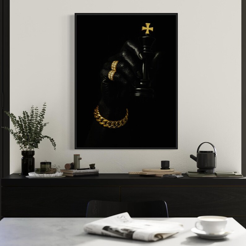 May include: A framed art print featuring a black chess king with a gold cross on top, held by a gloved hand adorned with gold rings and a gold chain bracelet. The background is solid black, creating a striking contrast.