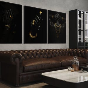 May include: Three framed black art prints with gold jewelry accents hang above a brown leather sofa. The prints depict hands and a torso adorned with gold jewelry. A marble-topped coffee table and a black cabinet complete the room.