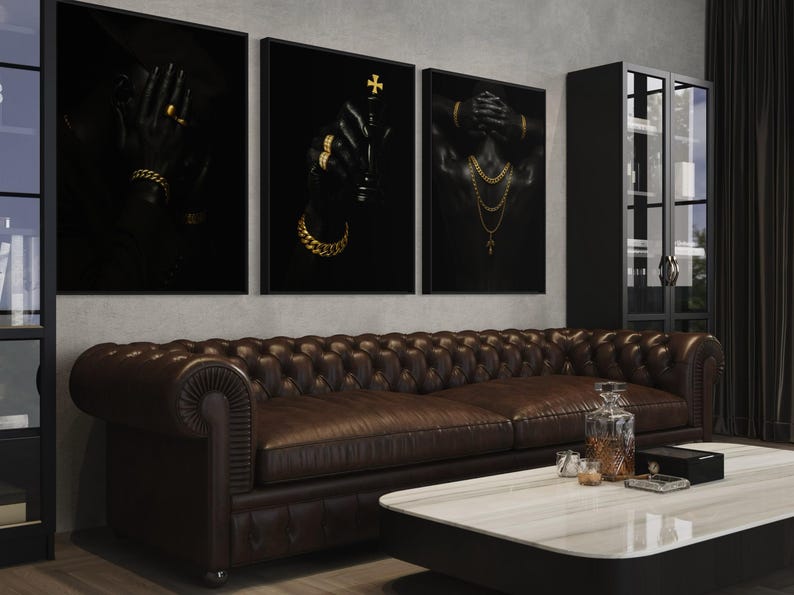May include: Three framed black art prints with gold jewelry accents hang above a brown leather sofa. The prints depict hands and a torso adorned with gold jewelry. A marble-topped coffee table and a black cabinet complete the room.