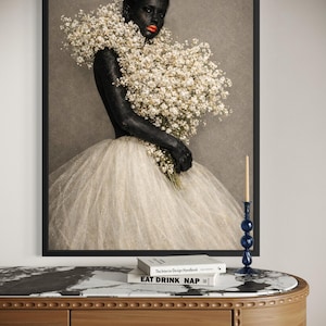 May include: A framed art print featuring a person with dark skin and red lipstick, adorned with a large bouquet of white flowers. The person wears a white tulle skirt. A blue candle holder and books are on a wooden table.