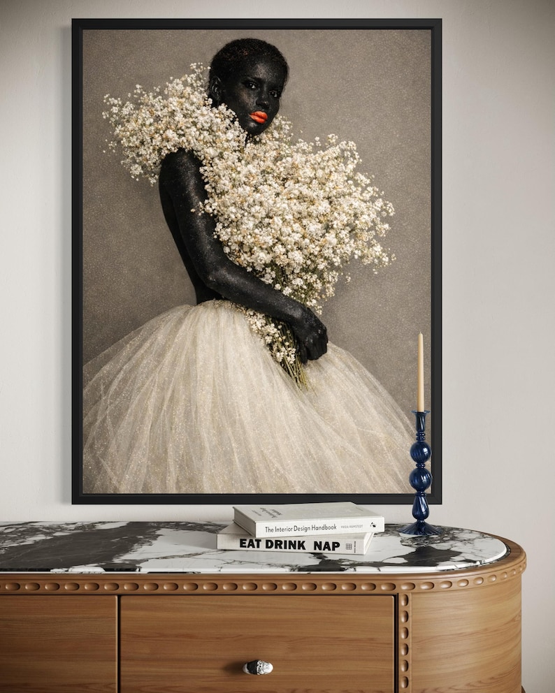May include: A framed art print featuring a person with dark skin and red lipstick, adorned with a large bouquet of white flowers. The person wears a white tulle skirt. A blue candle holder and books are on a wooden table.