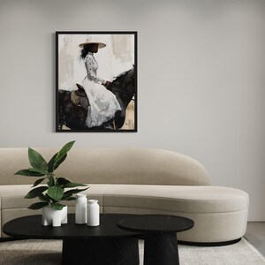 May include: A framed print shows a person on a black horse, wearing a white dress and a wide-brimmed hat. The background has neutral tones. The artwork is displayed above a beige sofa and black coffee table.
