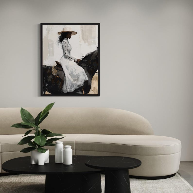 May include: A framed print shows a person on a black horse, wearing a white dress and a wide-brimmed hat. The background has neutral tones. The artwork is displayed above a beige sofa and black coffee table.