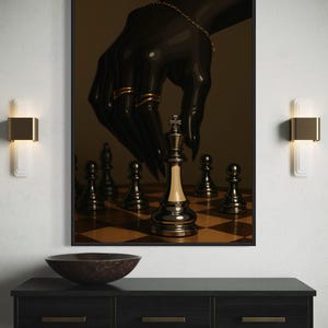 Op de afbeelding: Een ingelijste kunstdruk toont een zwarte hand met gouden ringen en een armband, boven een schaakbord. De koning schaakstuk staat centraal. Het kunstwerk wordt weergegeven boven een donkere houten console.