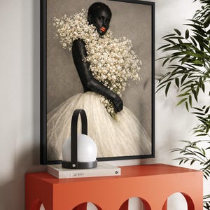 May include: A framed art print featuring a person with dark skin and bright red lipstick, adorned with white flowers. The person is wearing a white tulle dress. A modern lamp and a book are on an orange table.