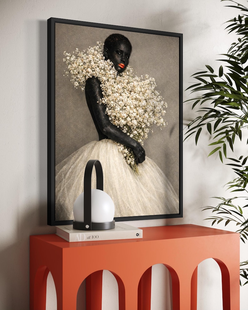 May include: A framed art print featuring a person with dark skin and bright red lipstick, adorned with white flowers. The person is wearing a white tulle dress. A modern lamp and a book are on an orange table.