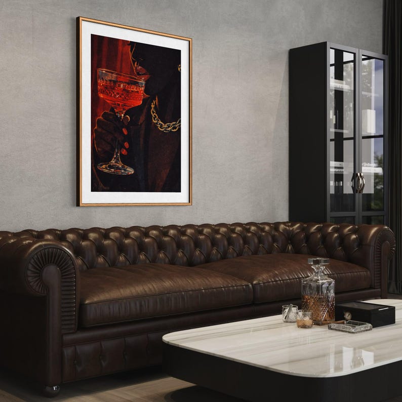 May include: A framed art print depicts a person holding a glass of red liquid, with a gold chain necklace. The artwork is set against a red backdrop. The print is in a gold frame, hanging above a brown leather sofa.