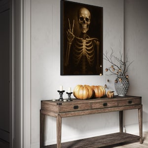Puede incluir: Una obra de arte enmarcada representa un esqueleto haciendo el signo de la paz. Una consola de madera exhibe calabazas, velas y un jarr&oacute;n decorativo con ramas. La escena evoca un tema de Halloween o oto&ntilde;o.