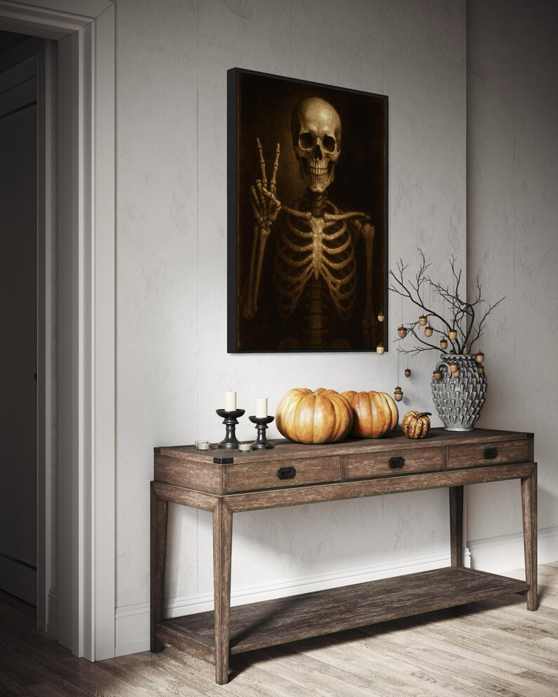 Puede incluir: Una obra de arte enmarcada representa un esqueleto haciendo el signo de la paz. Una consola de madera exhibe calabazas, velas y un jarr&oacute;n decorativo con ramas. La escena evoca un tema de Halloween o oto&ntilde;o.