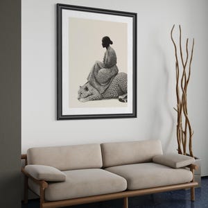 May include: A framed black and white art print depicts a person seated on a leopard. The artwork is displayed above a beige sofa with wooden frame. A tall, decorative plant in a gray pot stands to the right of the artwork.