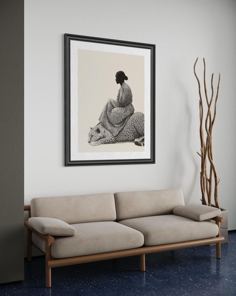May include: A framed black and white art print depicts a person seated on a leopard. The artwork is displayed above a beige sofa with wooden frame. A tall, decorative plant in a gray pot stands to the right of the artwork.